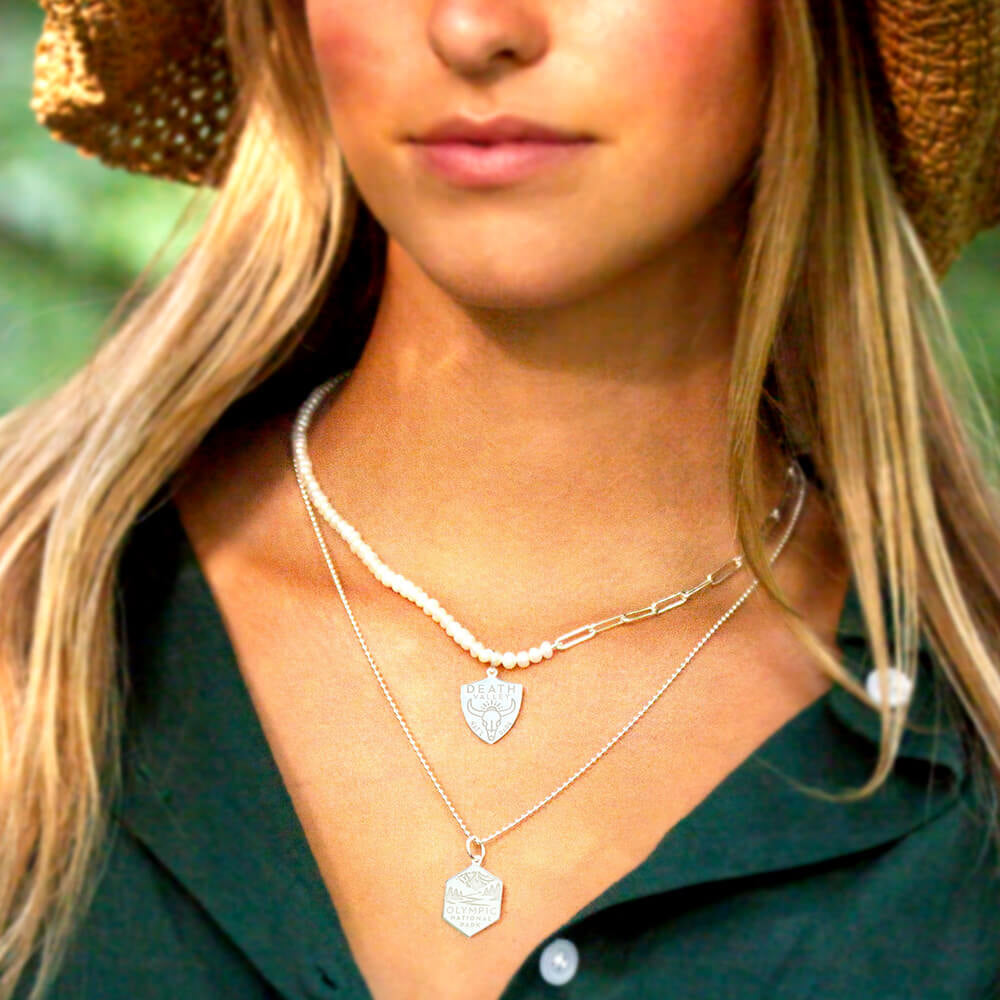 A woman is wearing two layered necklaces featuring National Park-themed charms. One necklace is a pearl paperclip chain with a charm representing Death Valley National Park, featuring an engraved design. The second necklace is a silver ball chain with a hexagonal charm representing Olympic National Park, also featuring an engraved design.
