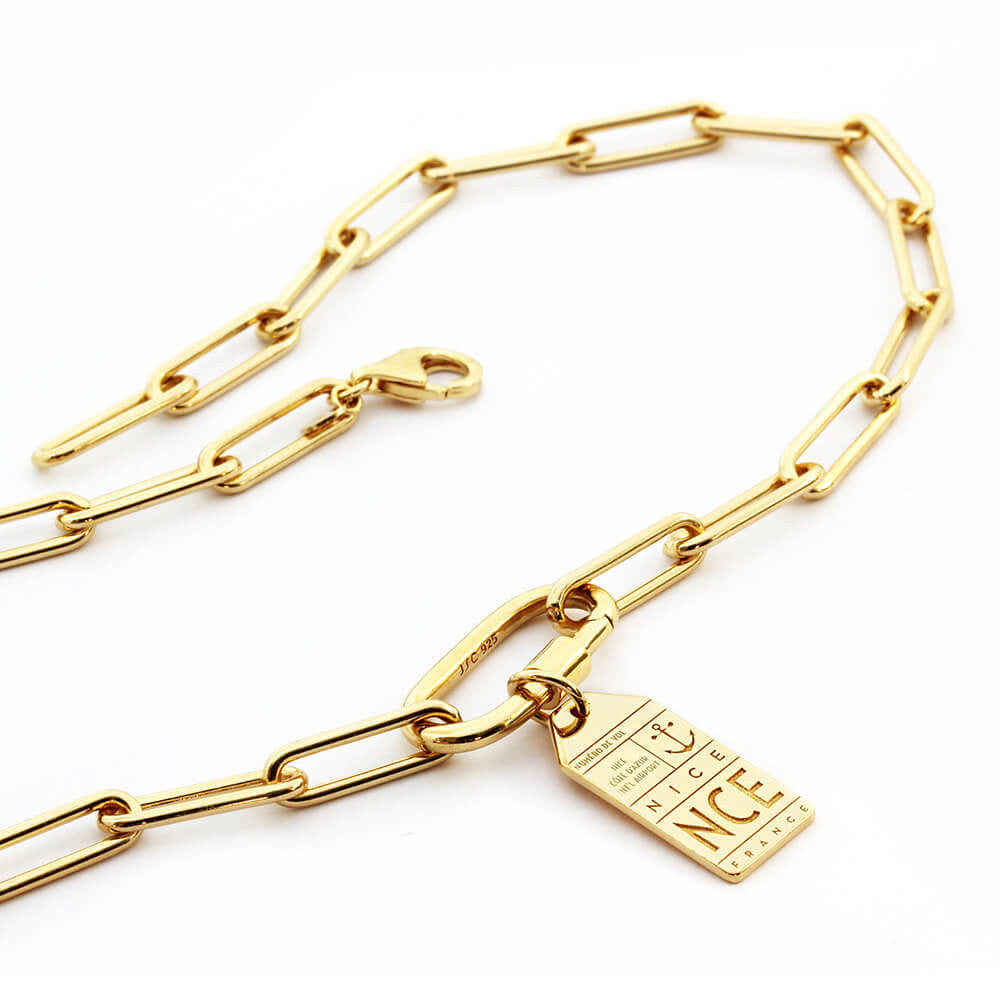 Photo featuring the Daily Charm Necklace in gold vermeil from Jet Set Candy. The bracelet features long rectangular links with rounded corners and an inch-long carabiner that is shown with a silver travel charm for Nice France with the airport code NCE. The bracelet is shown snaked on a white background.