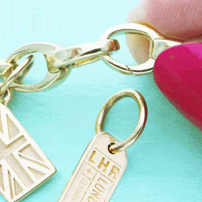A close up animated gif of hand with dark pink finger nails opening a link of the Solid 14k Gold Slim Infinity Link Charm Bracelet from Jet Set Candy and placing a solid gold London charm featuring LHR and a Union Jack engraved on it. The bracelet is being  The bracelet is photographed on a mini green background. The bracelet is made in Italy.