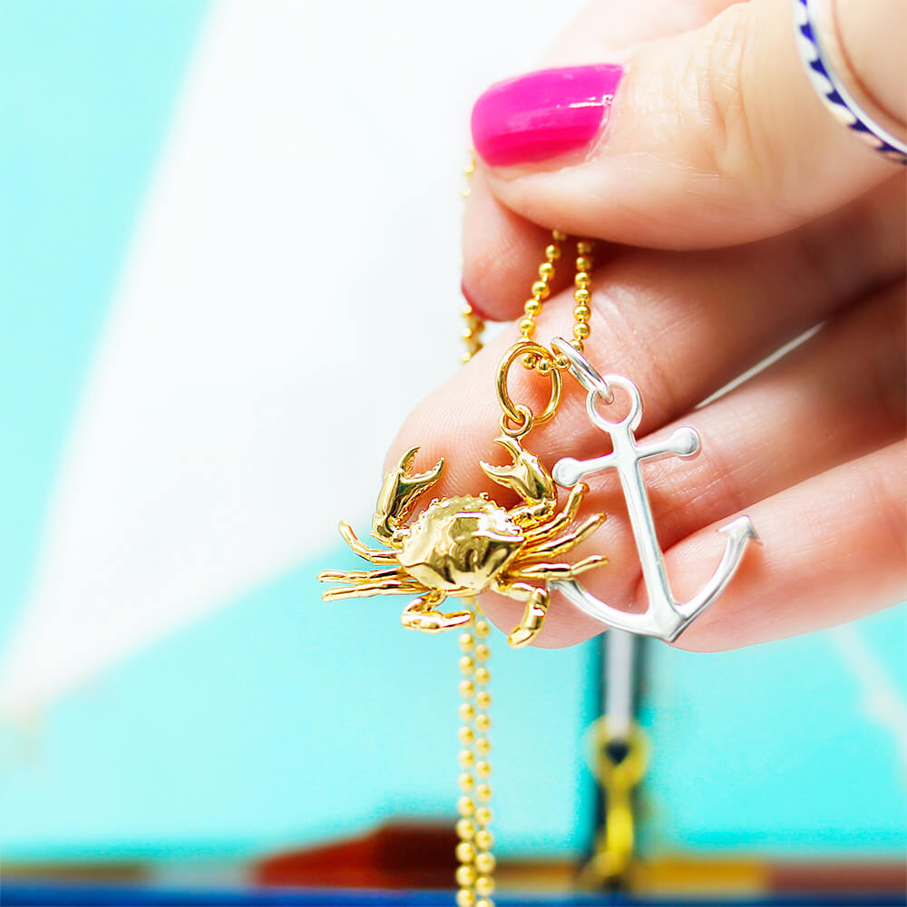 Gold crab and anchor charm by Jet Set Candy, held by model on a blurred background.