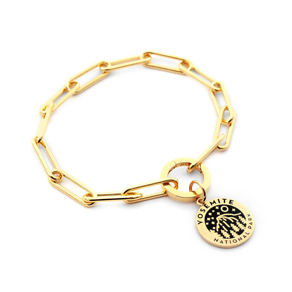 The image shows a gold vermeil 360 daily bracelet, a paperclip chain bracelet featuring a charm representing Yosemite National Park. The charm is round with a black enamel design of a landscape and starry sky, along with the text 