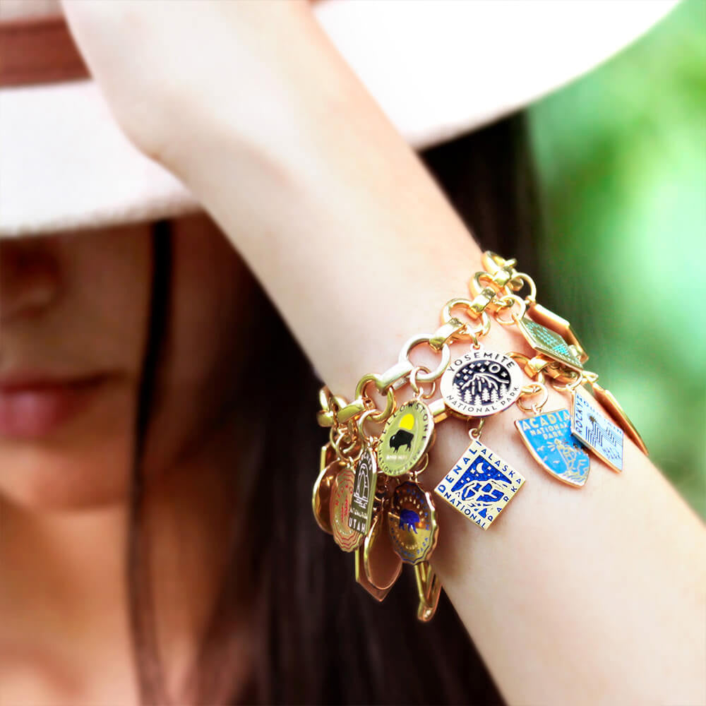 Gold vermeil infinity bracelet with opening links and enamel charms that represent parks such as Yosemite, Yellowstone, Acadia, Denali, and others. Each charm is detailed with unique engraved and enamel designs in various shapes, including circles, hexagons, and shields, and vibrant colors like blue, yellow, green, and black. 