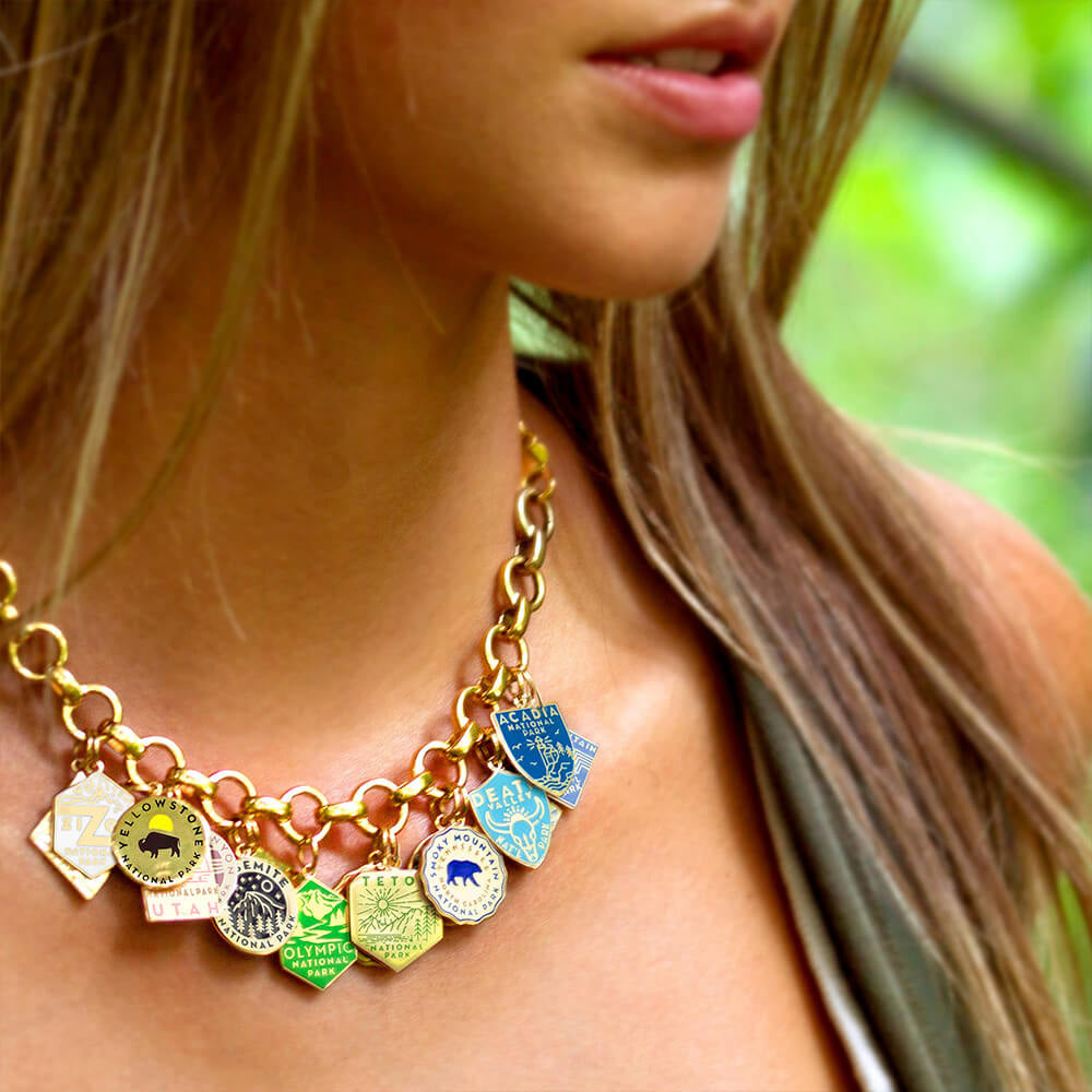A woman wearing a gold vermeil infinity necklace with opening individual links adorned with multiple National Park-themed charms. The charms include designs representing parks such as Zion, Yellowstone, Yosemite, Arches, Olympic, Teton, Smoky Mountain, Death Valley, Acadia, and Rocky Mountain. Each charm features unique engraved and enamel designs in various shapes, including hexagons, circles, and shields, with vibrant colors like blue, green, yellow