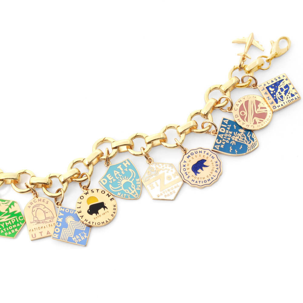 The image shows a gold vermeil or solid gold infinity bracelet with opening links with gold and enamel National Park-themed charms attached. Each charm represents a different park and is designed with distinct colors and graphics. Parks included are Arches, Olympic, Rocky Mountain, Yellowstone, Death Valley, Zion, Smoky Mountain, Acadia, Grand Canyon, and Denali. The charms are in various shapes such as shields, circles, hexagons, and diamonds, and feature colors like green, blue, yellow, pink, and beige