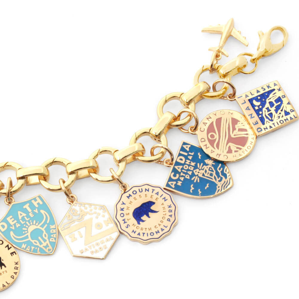 The image shows a gold bracelet with enamel National Park-themed charms attached. Each charm represents a different park and is designed with distinct colors and graphics. Parks included are Death Valley, Zion, Smoky Mountain, Acadia, Grand Canyon, and Denali. The charms are in various shapes such as shields, circles, hexagons, and diamonds, and feature colors like green, blue, yellow, pink, and beige. The bracelet has a clasp and a small airplane charm attached to it