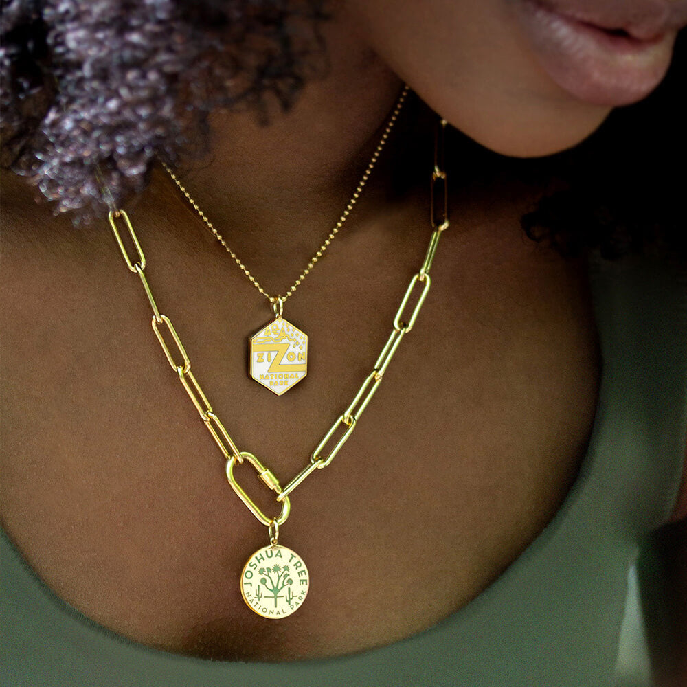 A person wearing two layered gold vermeil necklaces, each featuring a National Park-themed charm. The shorter necklace is a gold ball chain with a hexagonal charm representing Zion National Park, featuring an enamel design in white. The longer necklace is a gold paperclip chain with a carabiner and round charm representing Joshua Tree National Park, featuring an engraved design in green. 