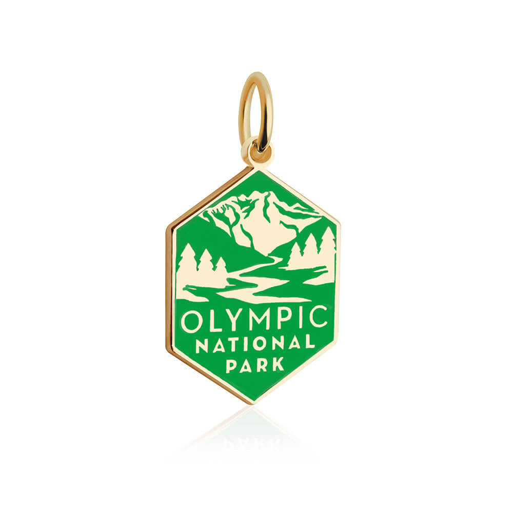 The image displays a hexagonal gold vermeil charm and a green enamel background. The charm features white enamel illustrations of a mountain range, evergreen trees, and a winding river. Below the illustration, the text 