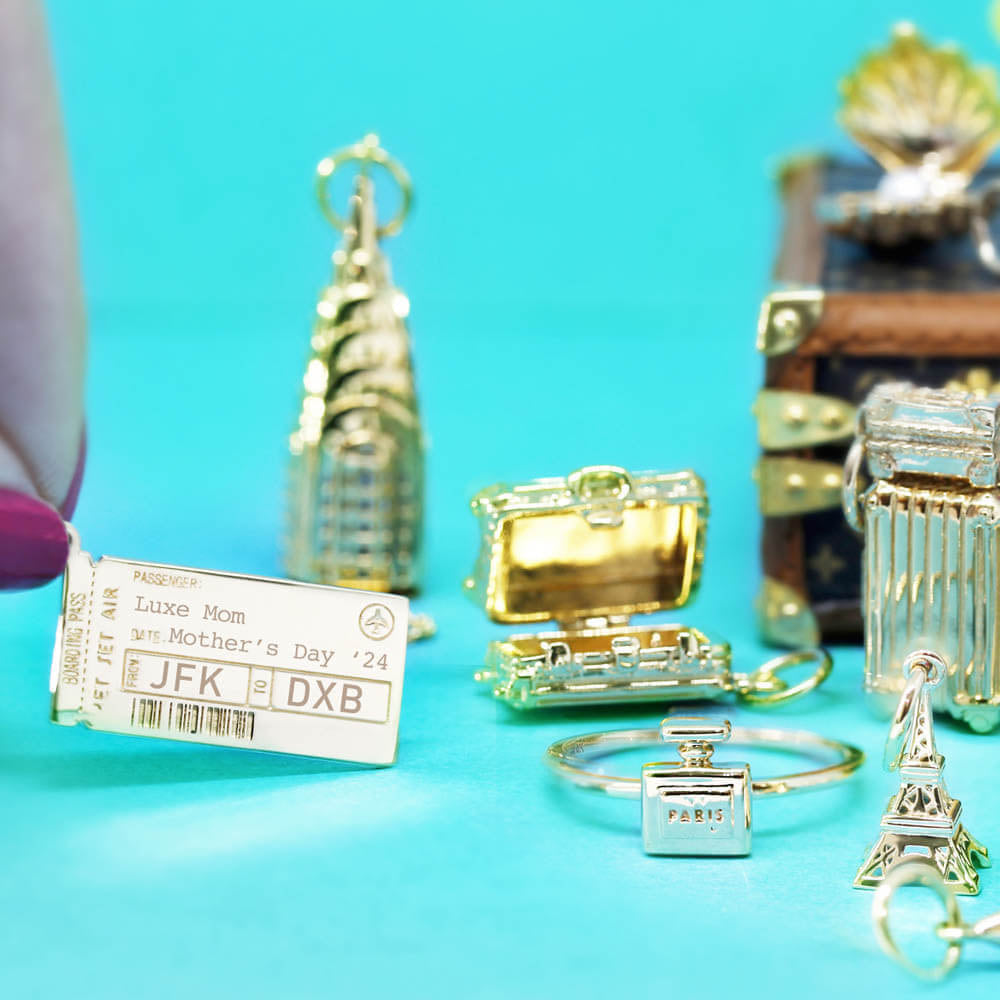 Gold boarding pass charm with Luxe Mom, JFK to DXB engravings, airplane icon, and barcode on a rectangular tag.