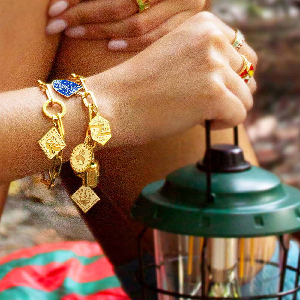 a person wearing two gold vermeil paperclip chain bracelets, each featuring multiple National Park-themed charms. The charms have engraved and enamel designs representing various parks, including Acadia, Zion, Yellowstone, Glacier, and Sequoia. The bracelets are layered on the wrist, creating a stylish and adventurous look.