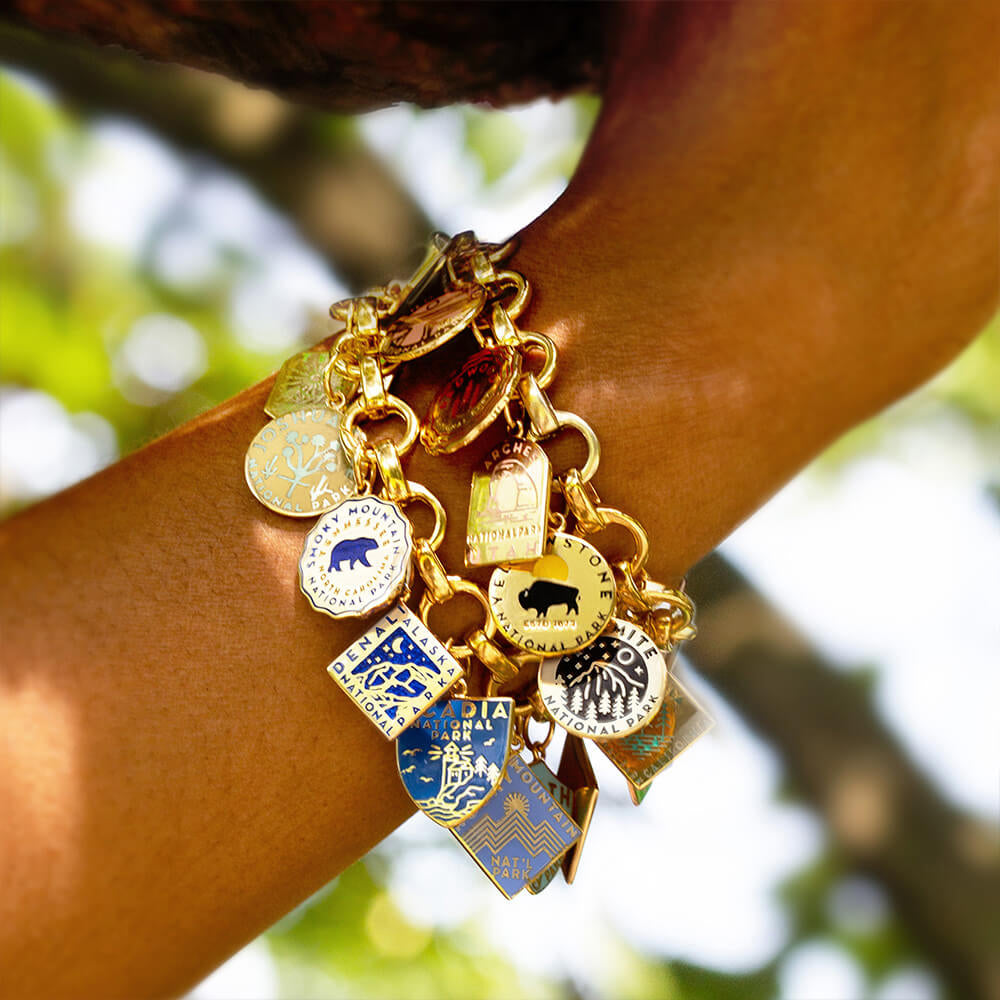 Gold vermeil infinity charm bracelets with individual opening links, each featuring a variety of National Park-themed charms. The charms represent different parks, including Smoky Mountain, Yellowstone, Denali, Acadia, Yosemite, Arches, and Rocky Mountain, among others. Each charm is detailed with unique engraved and enamel designs in various shapes such as circles, hexagons, and shields. The charms display vibrant enamel colors like blue, green, red, and yellow.