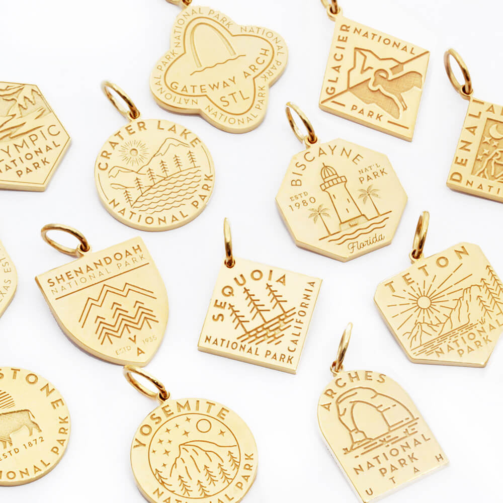 Gold vermeil and solid gold charms including designs representing parks such as Gateway Arch, Glacier, Olympic, Crater Lake, Biscayne, Denali, Shenandoah, Sequoia, Teton, Yellowstone, Yosemite, and Arches. Each charm features unique engraved designs in various shapes, including hexagons, circles, shields, and custom shapes.