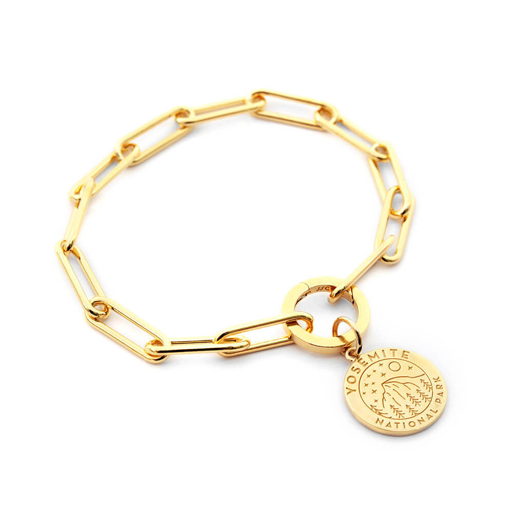 The image shows a gold vermeil 360 daily bracelet, a paperclip chain bracelet featuring a charm representing Yosemite National Park. The charm is round with an engraved design of a landscape and starry sky, along with the text 
