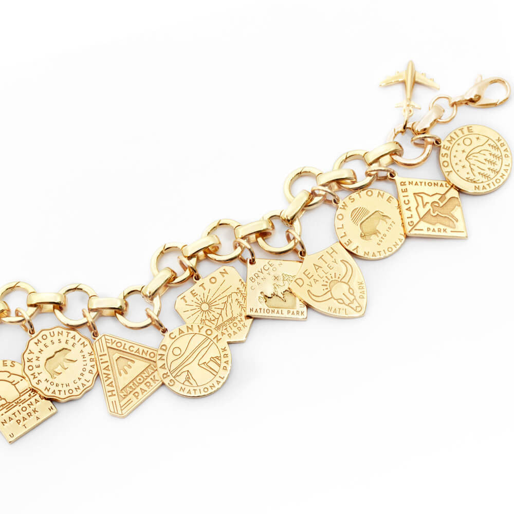The image shows a gold vermeil or solid gold infinity bracelet with open links with engraved National Park-themed charms attached. Each charm represents a different park and features unique engraved designs. The charms are in various shapes such as shields, circles, hexagons, and diamonds. Parks included are Smoky Mountain, Hawaii Volcanoes, Grand Canyon, Teton, Bryce Canyon, Death Valley, Yellowstone, Glacier, and Yosemite. The bracelet has a clasp and a small airplane charm attached to it