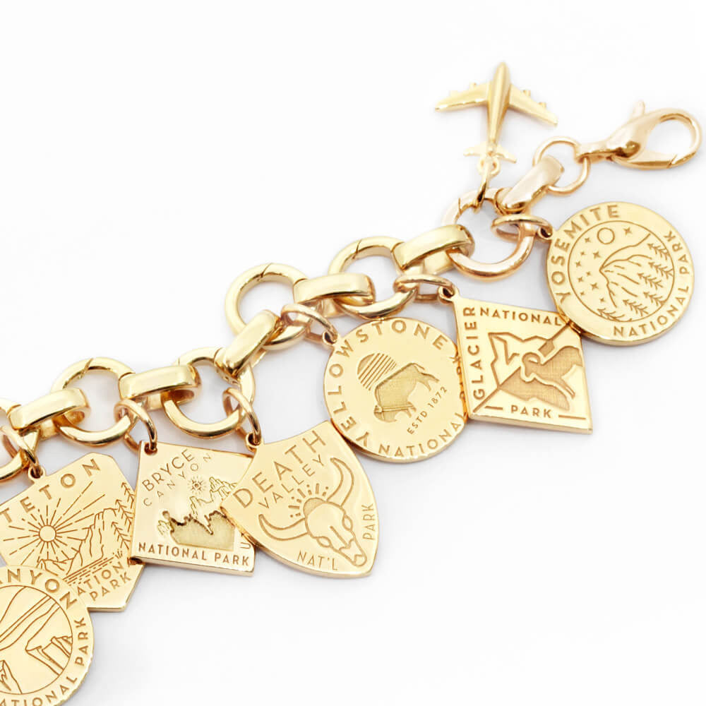 The image shows a gold bracelet with gold vermeil or solid gold National Park-themed charms attached. Each charm represents a different park and is designed with distinct colors and graphics. Parks included are Death Valley, Zion, Smoky Mountain, Acadia, Grand Canyon, and Denali. The charms are in various shapes such as shields, circles, hexagons, and diamonds, and feature colors like green, blue, yellow, pink, and beige. The bracelet has a clasp and a small airplane charm attached to it.