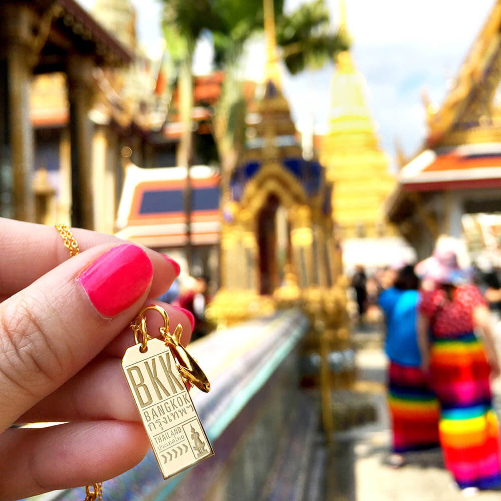 Gold luggage tag charm featuring BKK airport code, Bangkok Thai text, airplane motif, and traditional temple statue engraving.