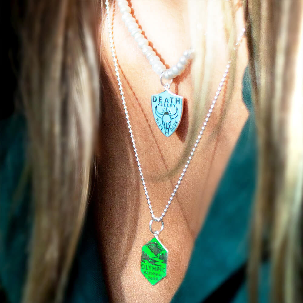  A woman is wearing two layered necklaces featuring National Park-themed charms. One necklace is a pearl paperclip chain with a charm representing Death Valley National Park, featuring an engraved design. The second necklace is a silver ball chain with a hexagonal charm representing Olympic National Park, also featuring an engraved design. 