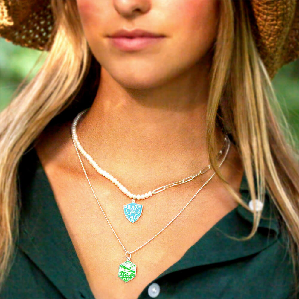  A woman is wearing two layered necklaces featuring National Park-themed charms. One necklace is a pearl paperclip chain with a charm representing Death Valley National Park, featuring an engraved design. The second necklace is a silver ball chain with a hexagonal charm representing Olympic National Park, also featuring an engraved design. 