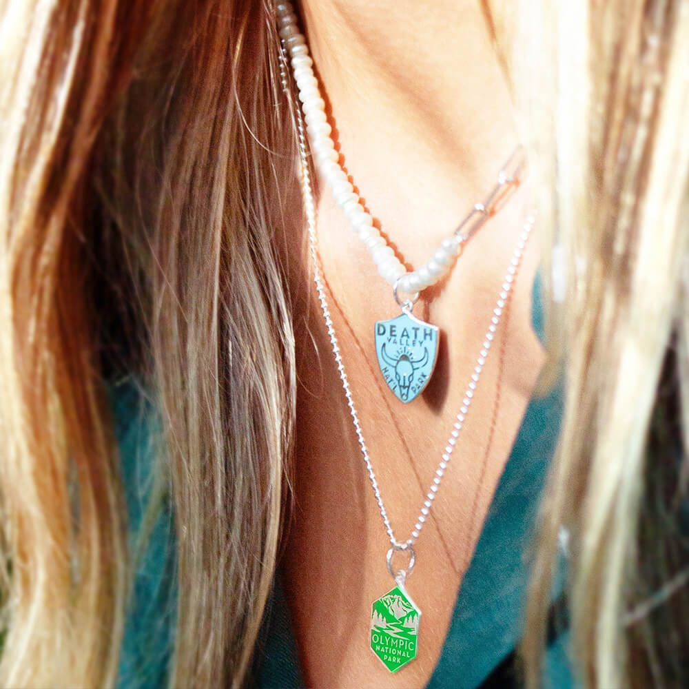  A woman is wearing two layered necklaces featuring National Park-themed charms. One necklace is a pearl paperclip chain with a charm representing Death Valley National Park, featuring an engraved design. The second necklace is a silver ball chain with a hexagonal charm representing Olympic National Park, also featuring an engraved design. 