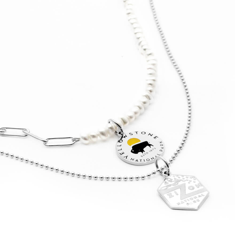 The image shows a set of two necklaces featuring National Park-themed charms. One is a pearl paperclip sterling silver necklace with a sterling silver charm representing Yellowstone National Park, featuring an engraved design of a bison and a yellow sun. The second necklace is a sterling silver ball chain with a hexagonal sterling silver charm representing Zion National Park, featuring enamel park graphics. The combination of pearl and silver chains gives a layered look