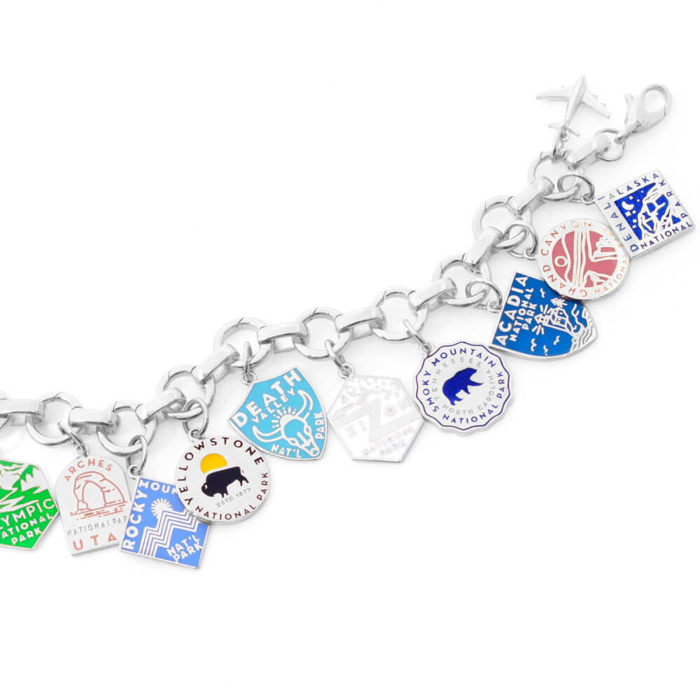 The image shows a sterling silver bracelet with enamel National Park-themed charms attached. Each charm represents a different park and is designed with distinct colors and graphics. Parks included are Arches, Olympic, Rocky Mountain, Yellowstone, Death Valley, Zion, Smoky Mountain, Acadia, Grand Canyon, and Denali. The charms are in various shapes such as shields, circles, hexagons, and diamonds, and feature colors like green, blue, yellow, pink, and beige