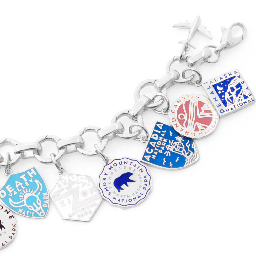  The image shows a sterling silver bracelet with enamel National Park-themed charms attached. Each charm represents a different park and is designed with distinct colors and graphics. Parks included are Death Valley, Zion, Smoky Mountain, Acadia, Grand Canyon, and Denali. The charms are in various shapes such as shields, circles, hexagons, and diamonds, and feature colors like green, blue, yellow, pink, and beige