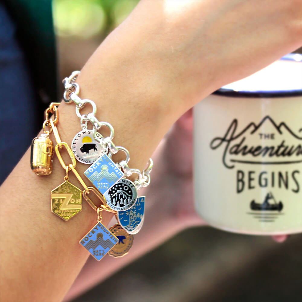 A sterling silver infinity bracelet with individual opening links adorned with various national park charms, including Yellowstone, Rocky Mountain, Yosemite, and a gold vermeil clip bracelet with Xion and Rocky Mountain enamel charms.