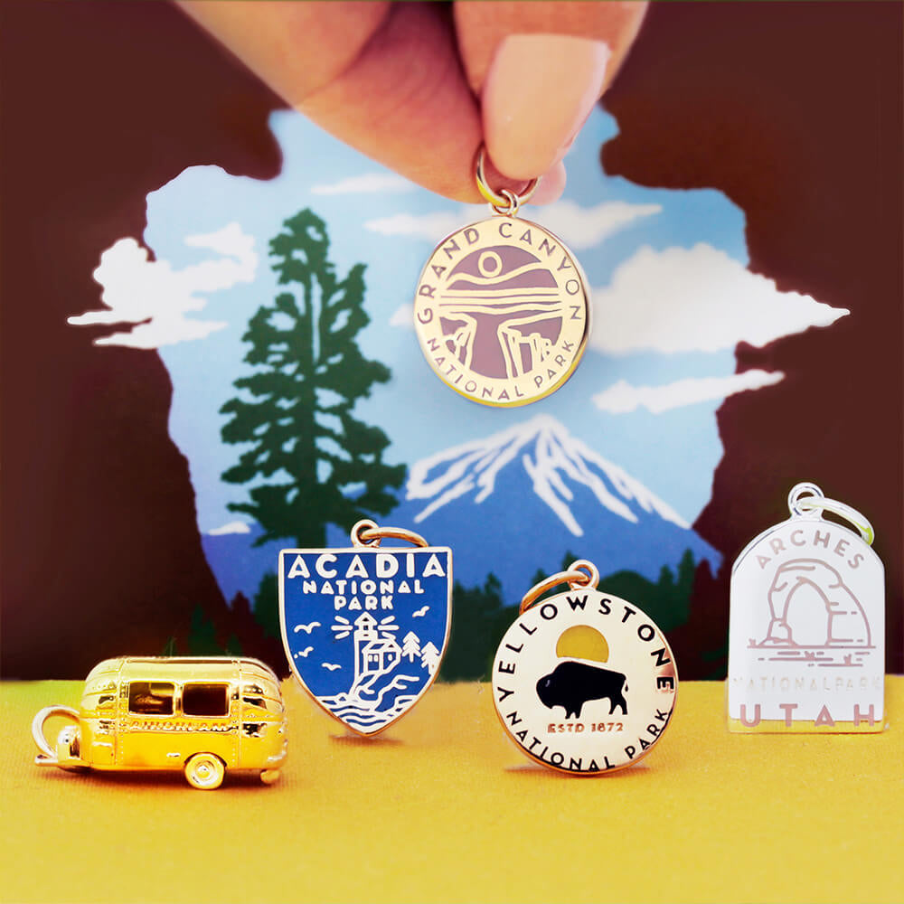 Collection of various National Park-themed charms set against a scenic background. The charms include designs representing parks such as Grand Canyon, Acadia, Yellowstone, and Arches, along with a custom-shaped gold vermeil trailer charm. Each charm features unique engraved and enamel designs in various shapes, including circles and shields.