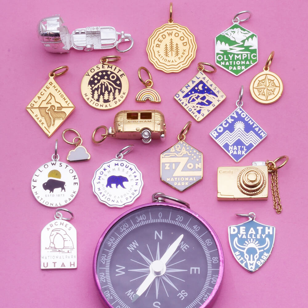 Collection of various National Park-themed charms on a pink background. The charms include designs representing parks such as Redwood, Olympic, Glacier, Yosemite, Denali, Rocky Mountain, Zion, Yellowstone, Smoky Mountain, Arches, and Death Valley. Each charm features unique engraved and enamel designs, with shapes including hexagons, circles, diamonds, and custom shapes like a trailer and a camera. 