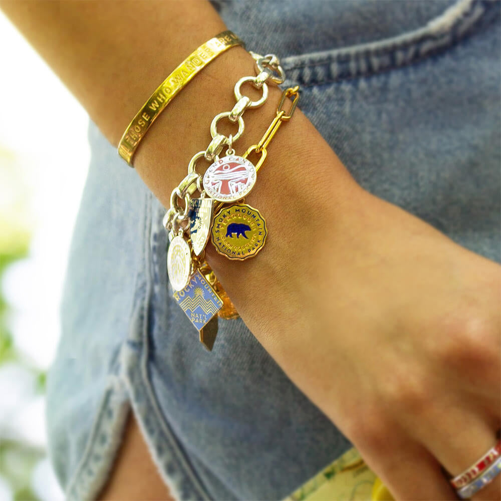 Cuff gold vermeil bracelet with travel quote, sterling silver infinity link charm bracelet with charms hanging representing national parks such as Grand Canyon, Smoky Mountain, Rocky Mountain, and others. Each charm features unique engraved and enamel designs in various shapes, including circles, hexagons, and shields, with colors like blue, red, and gold. 