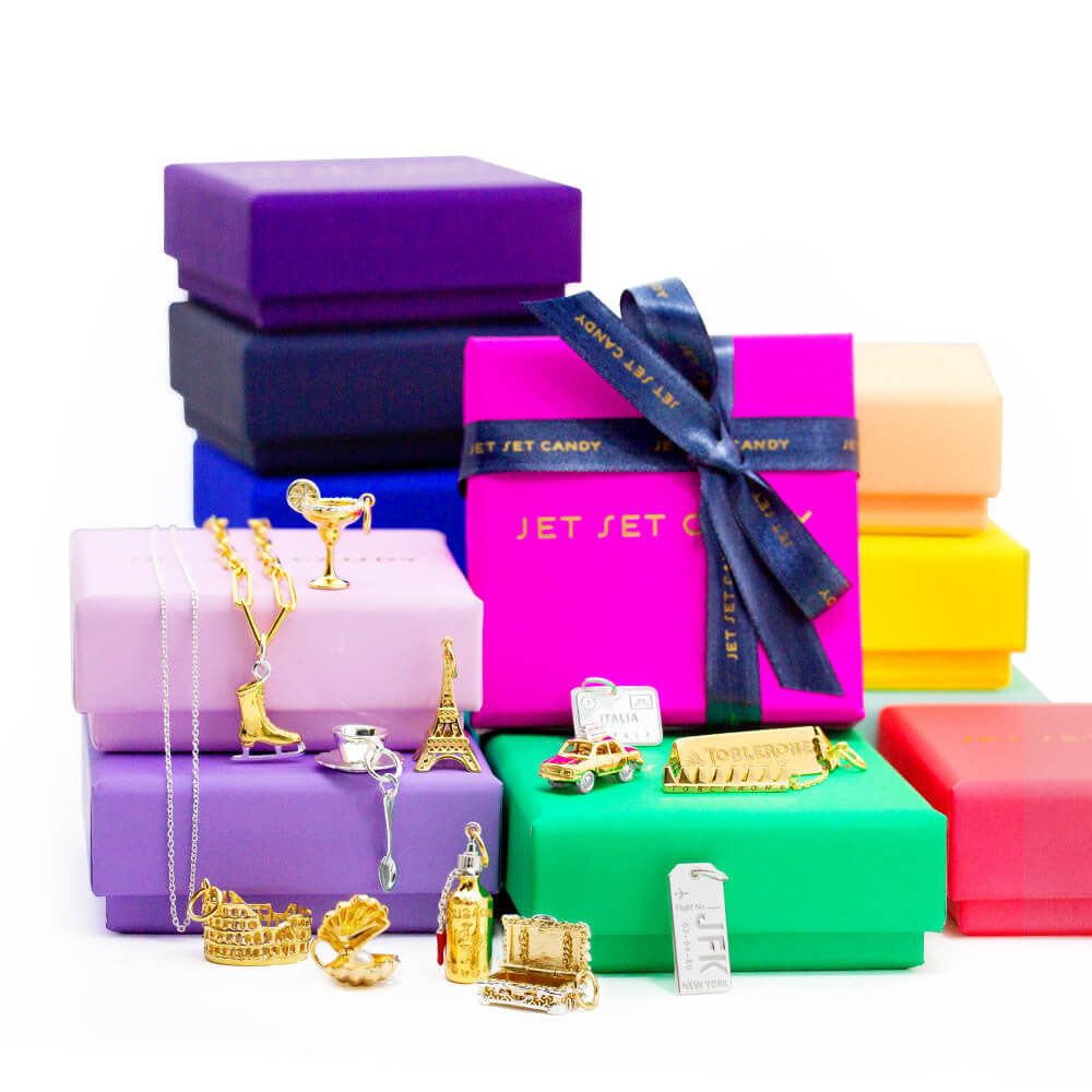 Gold and Sterling Silver charms featuring Eiffel Tower, Colosseum, JFK tag, and cocktail motifs displayed on colorful jewelry boxes.