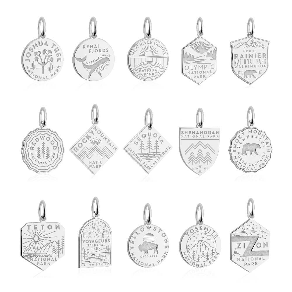 15 sterling silver National Park-themed charms, each representing a different park. The charms are arranged in three rows of five. Each charm has unique engraved designs and shapes, including shields, circles, hexagons, and diamonds. Parks represented include Joshua Tree, Kenai Fjords, New River Gorge, Olympic, Mount Rainier, Redwood, Rocky Mountain, Sequoia, Shenandoah, Smoky Mountain, Teton, Voyageurs, Yellowstone, Yosemite, and Zion.