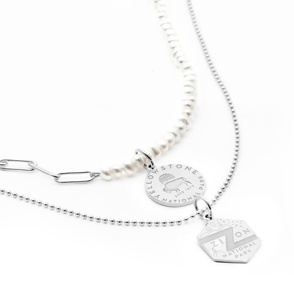 The image shows a set of two necklaces featuring National Park-themed charms. One is a pearl paperclip sterling silver necklace with a sterling silver charm representing Yellowstone National Park, featuring an engraved design of a bison and a sun. The second necklace is a sterling silver ball chain with a hexagonal gold vermeil charm representing Zion National Park, featuring engraved park graphics. The combination of pearl and sterling silver chains gives a layered look