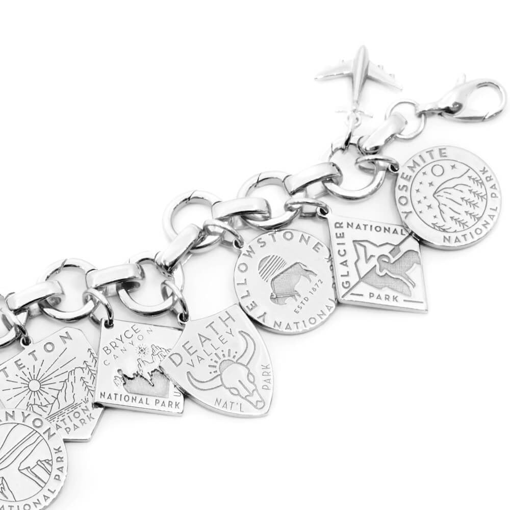 The image shows a sterling silver infinity bracelet with National Park-themed charms attached. Each charm represents a different park and is designed with different graphics. Parks included are Death Valley, Zion, Smoky Mountain, Acadia, Grand Canyon, and Denali. The charms are in various shapes such as shields, circles, hexagons, and diamonds, and feature colors like green, blue, yellow, pink, and beige. The bracelet has a clasp and a small airplane charm attached to it