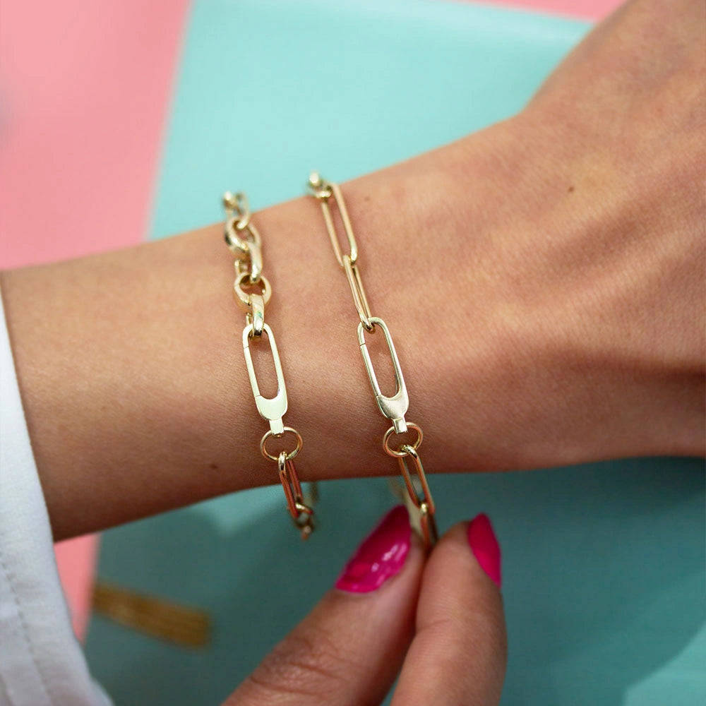 Gold Charm bracelets on a wrist featuring polished paperclip links and a distinctive elongated hook clasp design elements.