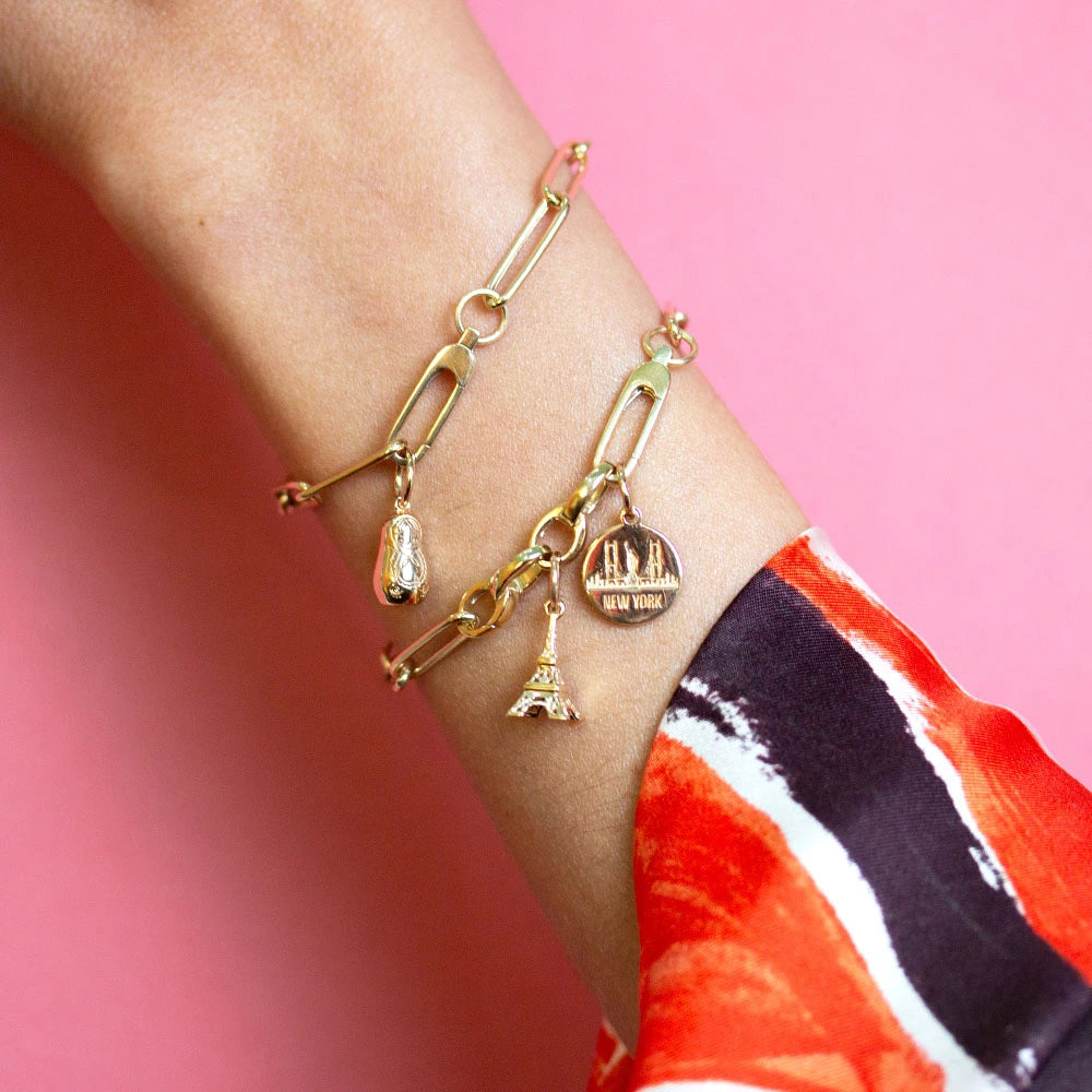 Gold charms including a 3D Eiffel Tower, round New York skyline disc, and matryoshka doll on paperclip link bracelets.