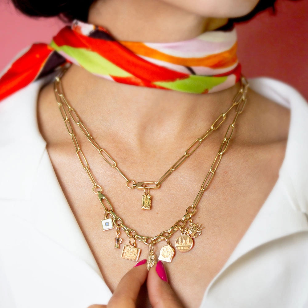 Gold charm necklace featuring various travel-themed pendants including a miniature passport, city skylines, and textured 3D luggage tag icons.
