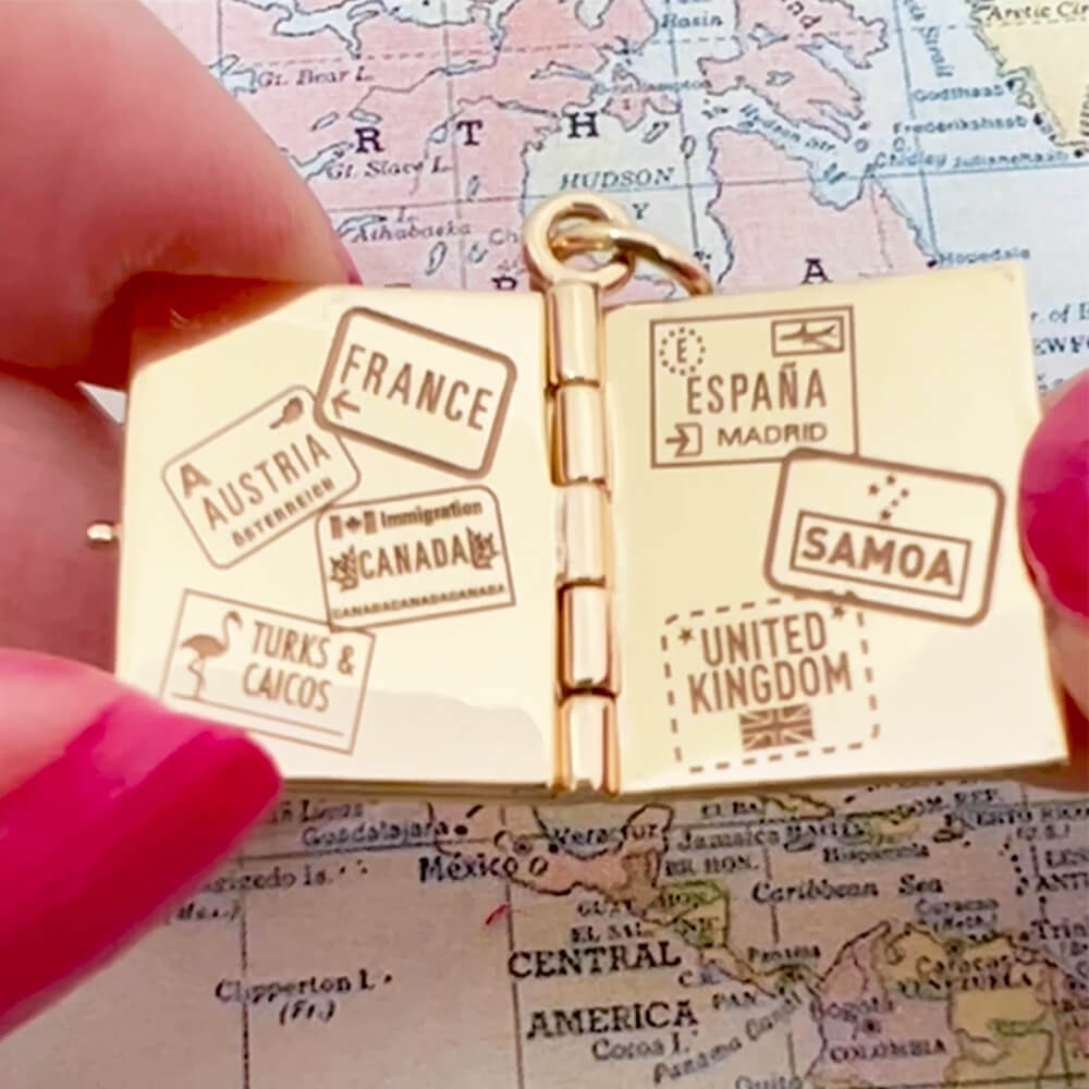 Bespoke Solid 14k Gold USA Passport Book Charm with blue enamel shown hanging on a chain in front of an airplane window overlooking an airplane wing and blue skies. background. This charm is fully customizable and you can choose up to 12 passport stamps to include on the solid gold pages inside. This charm is a one-of-a-kind souvenir.