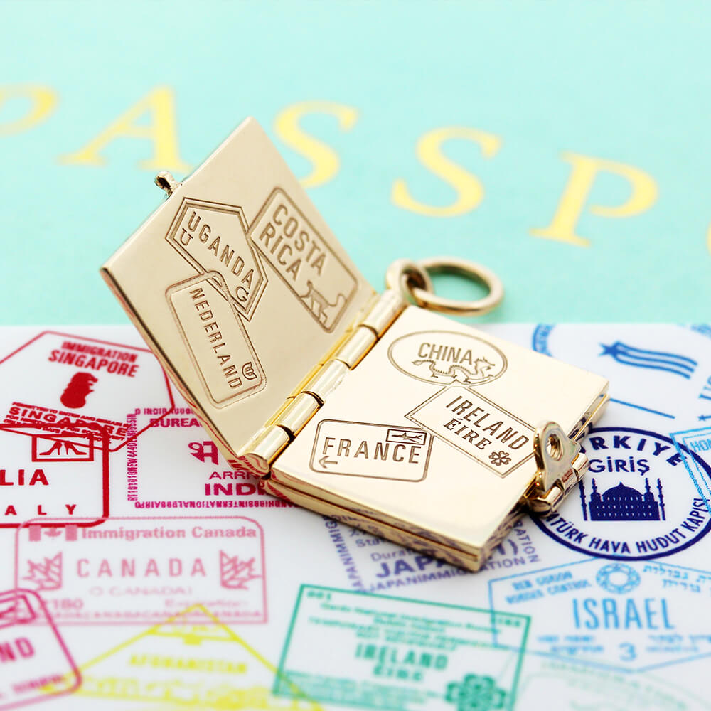 Bespoke Solid 14k Gold USA Passport Book Charm with blue enamel shown hanging on a chain in front of an airplane window overlooking an airplane wing and blue skies. background. This charm is fully customizable and you can choose up to 12 passport stamps to include on the solid gold pages inside. This charm is a one-of-a-kind souvenir.