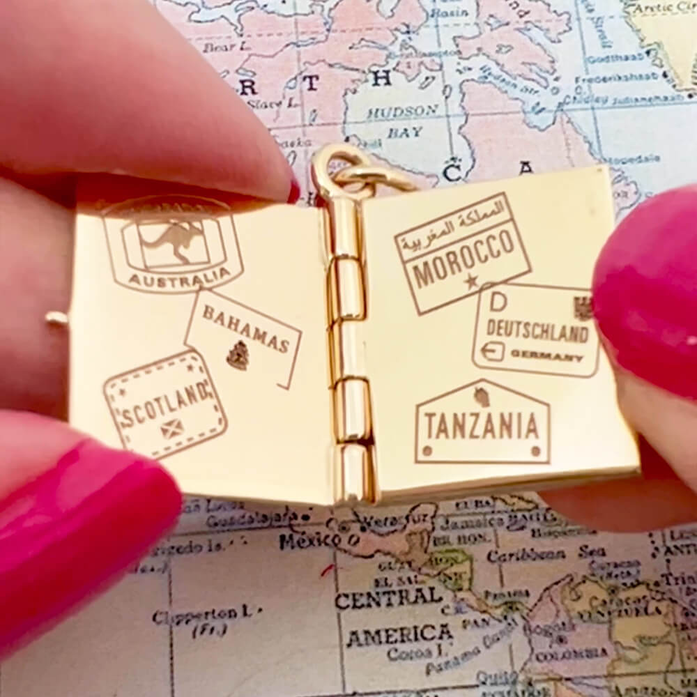 Bespoke Solid 14k Gold USA Passport Book Charm with blue enamel shown hanging on a chain in front of an airplane window overlooking an airplane wing and blue skies. background. This charm is fully customizable and you can choose up to 12 passport stamps to include on the solid gold pages inside. This charm is a one-of-a-kind souvenir.