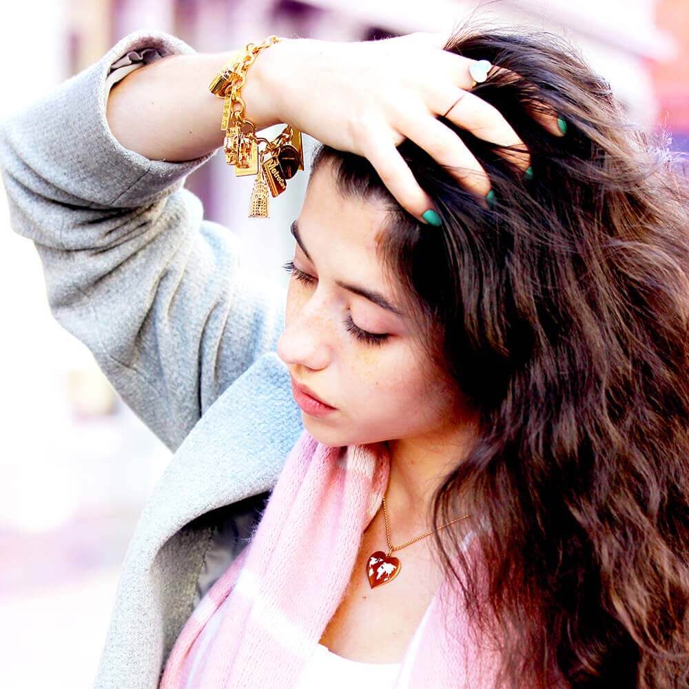 Brunette woman running fingers through her hair wearing a Gold Infinity Link Charm Bracelet from JET SET CANDY with a variety of gold New York-themed charms including a gold MetroCard Charm, a gold Chrysler Charm, and a gold NYC taxi charm. Woman is also wearing a gold Heart charm with a white enamel world map inside of it. All of the jewelry shown is from Jet Set Candy