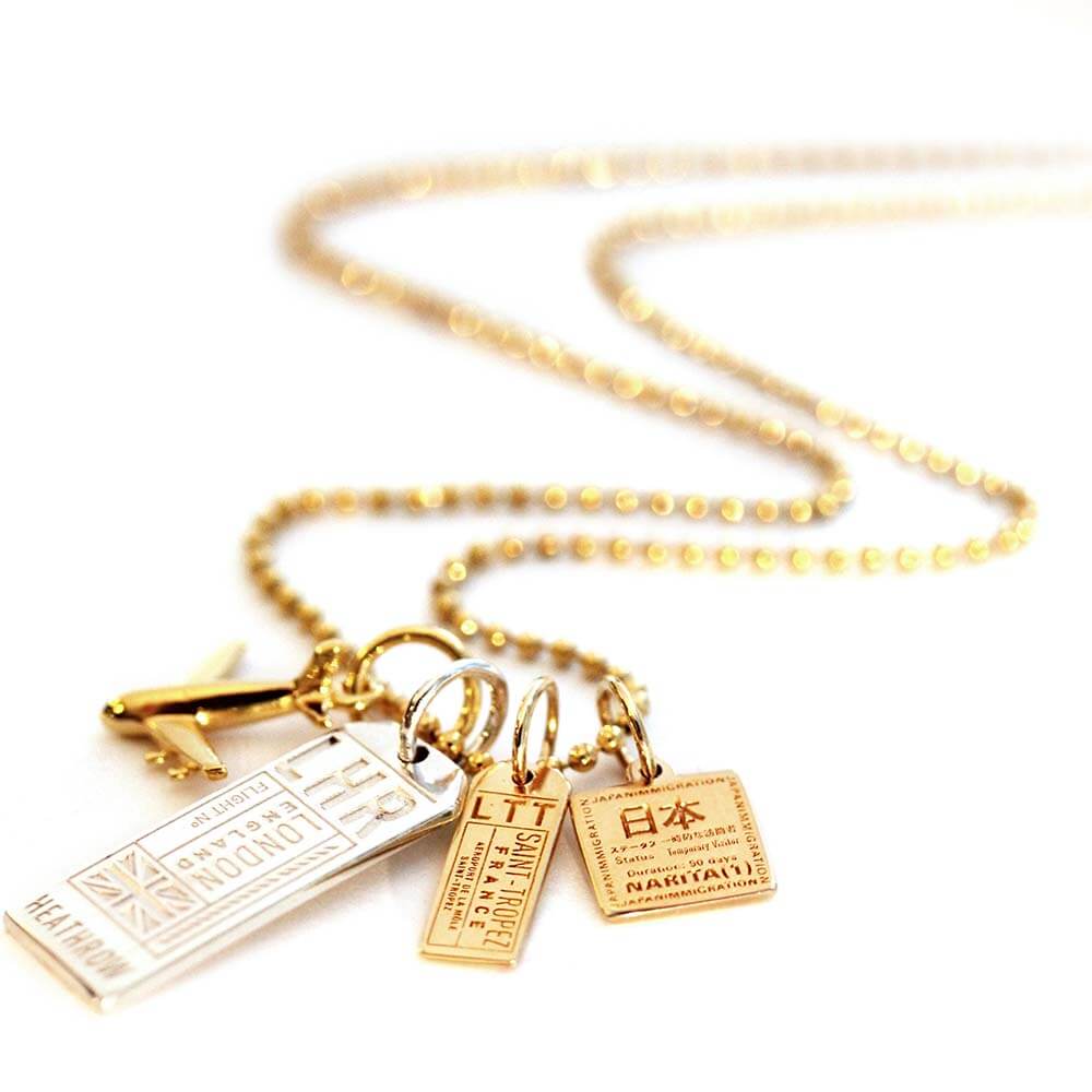  Charm necklace from Jet Set Candy featuring a gold plane charm, a silver LHR Luggage Tag Charm, and solid gold mini travel charms for St. Tropez LTT and a solid gold mini Japan passport stamp charm from Jet Set Candy. The chain is a gold ball chain and the necklace was photographed on a white background