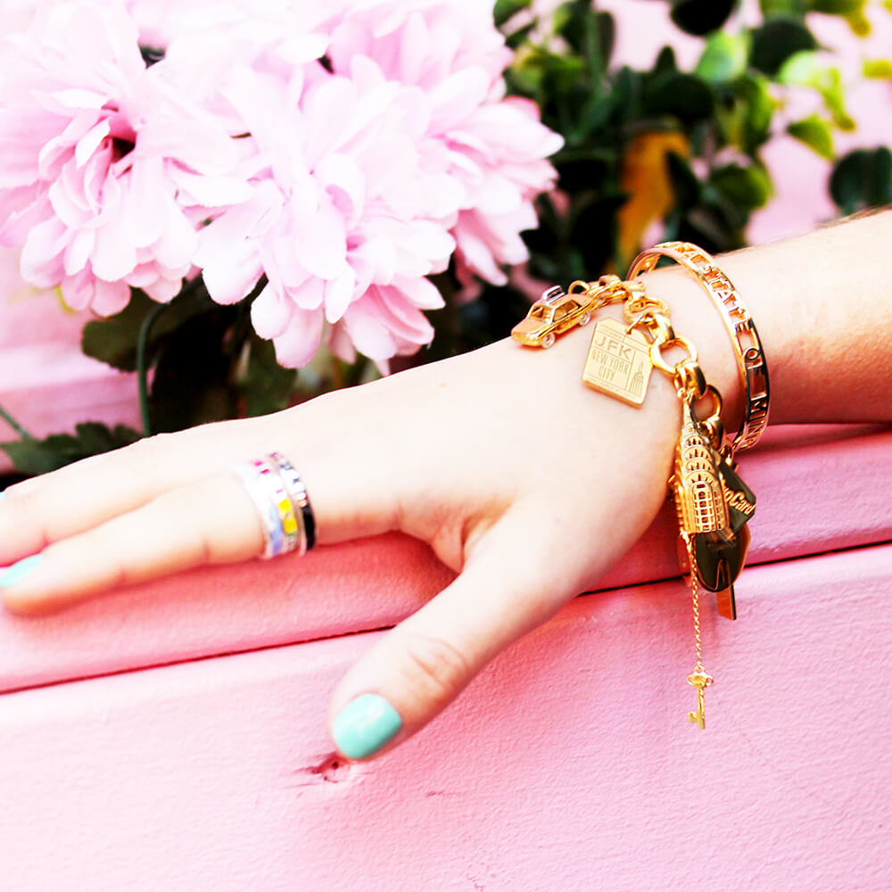Gold Infinity Link Charm Bracelet from Jet Set Candy featuring travel charms including a gold NYC Taxi charm, a gold JFK Luggage Tag Charm, and a gold Chrysler Building charm. The hand also shows a stack of rings on the index finger and a gold cuff that says 