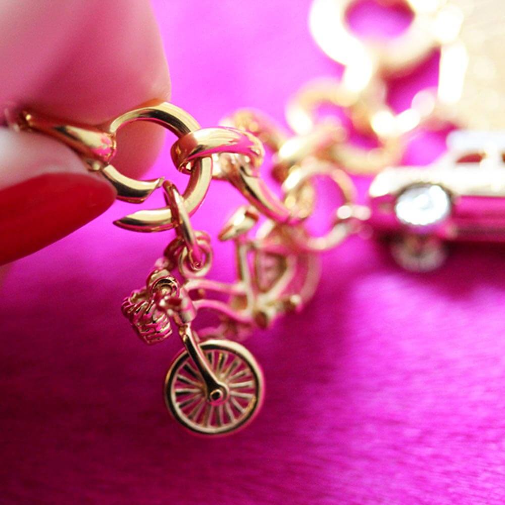 Close up of a finger with red nail polishing pushing in a link on a Gold Infinity Link Charm Bracelet from  JET SET CANDY and adding a gold bicycle charm to it. The bracelet is shown on a textured hot pink background.