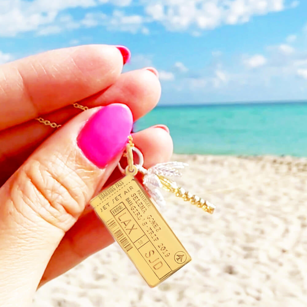 Photo of a solid 14k gold Boarding Pass Charm from the jewelry company Jet Set Candy. The charm is customizable and this one shows the name “Selena Gomez” engraved along with the airport codes “LAX” and “SJD” as well as a date. The photo was taken on a beach being held by a hand with hot pink nails. There is also a silver and gold palm tree charm being featured.