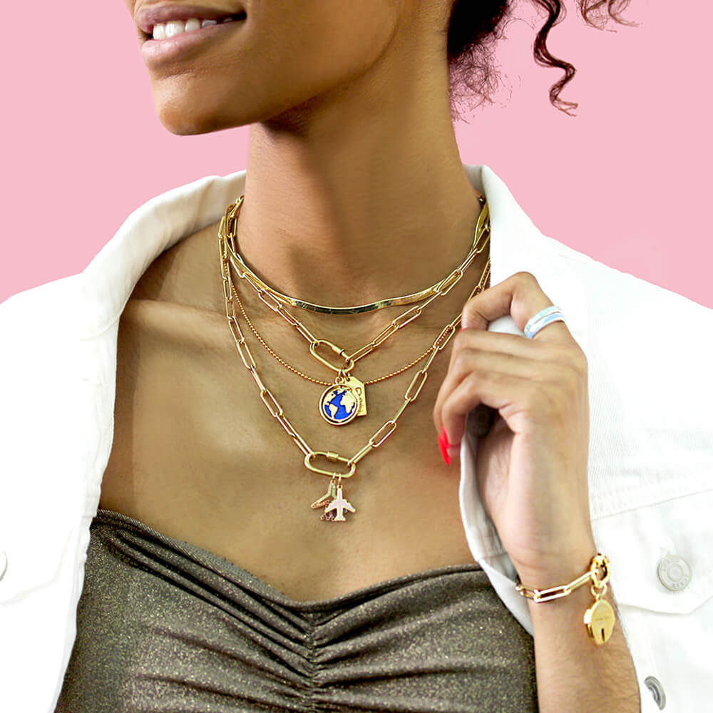 Photo of a model wearing a gold top, white jacket and several gold chains including gold Daily Charm Necklaces from Jet Set Candy. The gold chain necklaces feature long rectangular links with rounded corners, each with an inch-long carabiner charm clip. One of the charm clips is holding a gold and blue enamel world globe charm hanging from it and another has two airplane charms. The model is standing in front of a pale pink background.
