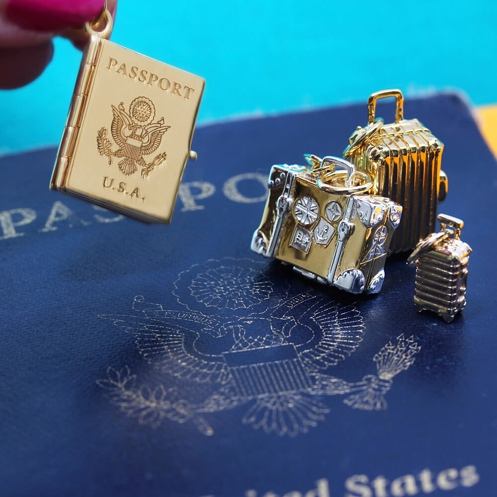 Hand with pink nail polish holding a Bespoke Solid 14k Gold USA Passport Book Charm  in front of an actual passport book as the background. This charm is fully customizable and you can choose up to 12 passport stamps to include on the solid gold pages inside. This charm is a one-of-a-kind souvenir. There are several other travel charms shown including 3 different gold suitcases charms.