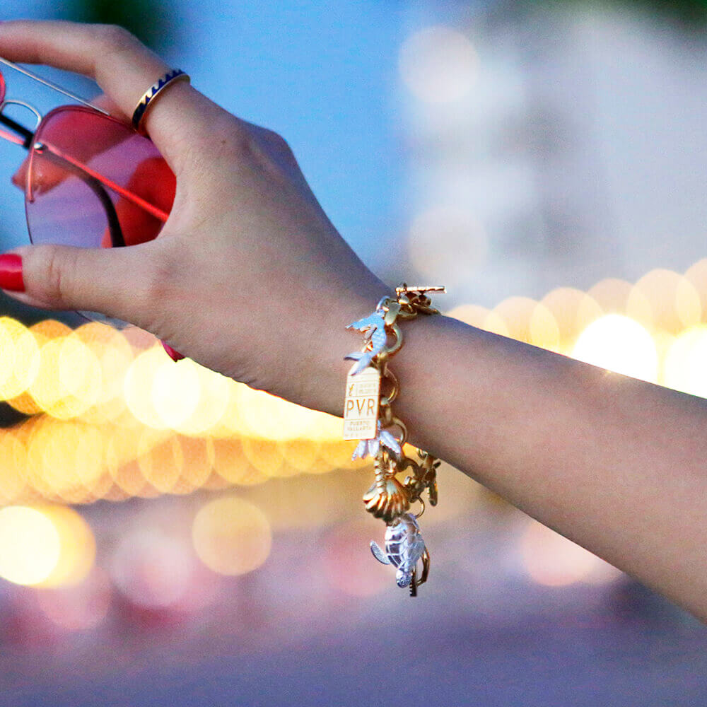 Hand holding pink glasses and wearing a blue and gold wave ring on their index finger and a Gold Infinity Link Charm Bracelet on their wrist with a silver mermaid charm, a gold PVR Puerto Vallarta Charm, a gold clamshell charm, and a gold and silver palm tree charm. The background features a nightscape with  out of focus lights