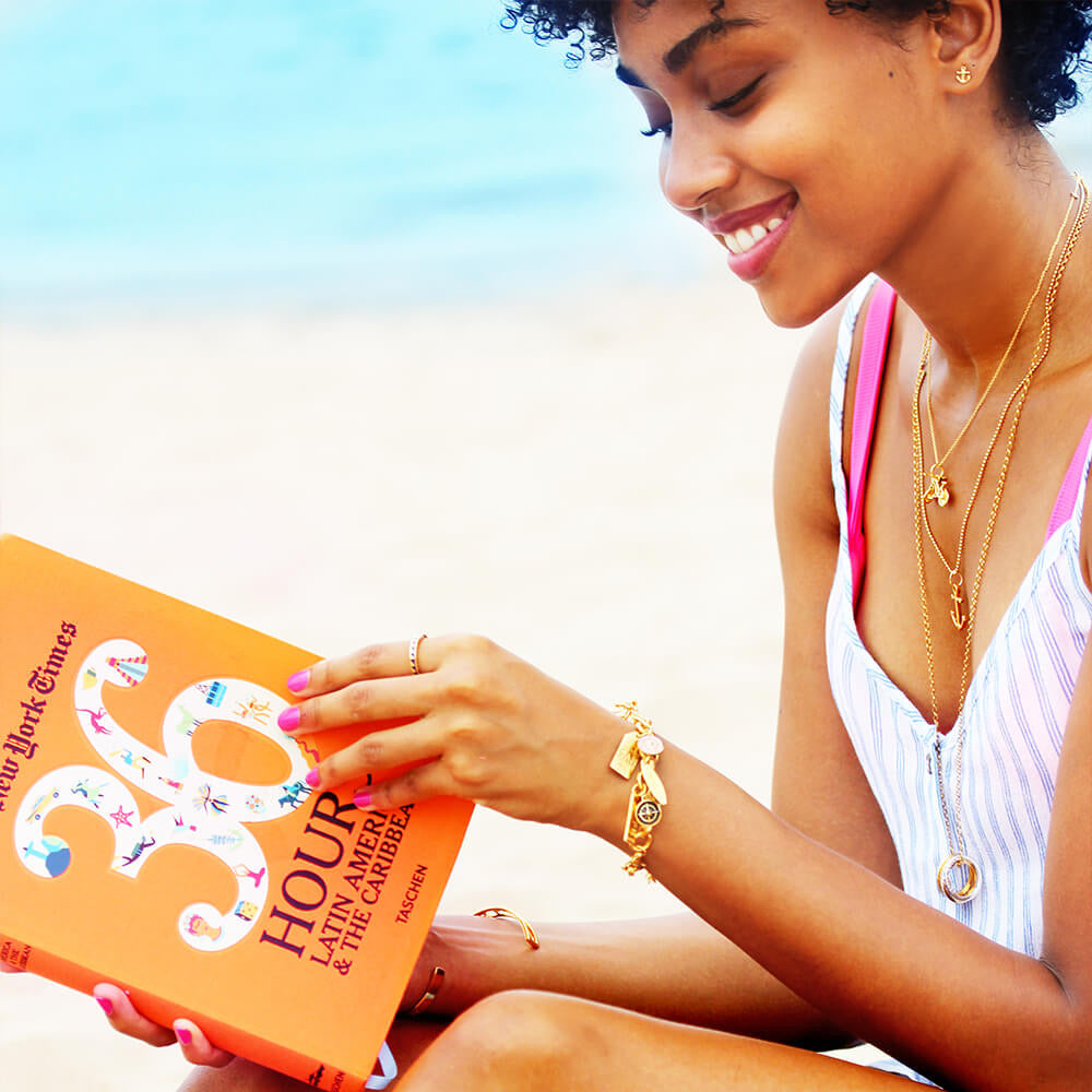 Woman on a beach reading an orange book titled “36 Hours in Latin America & the Caribbean from the New York Times and wearing a Solid Gold Infinity Link Charm Bracelet from JET SET CANDY.