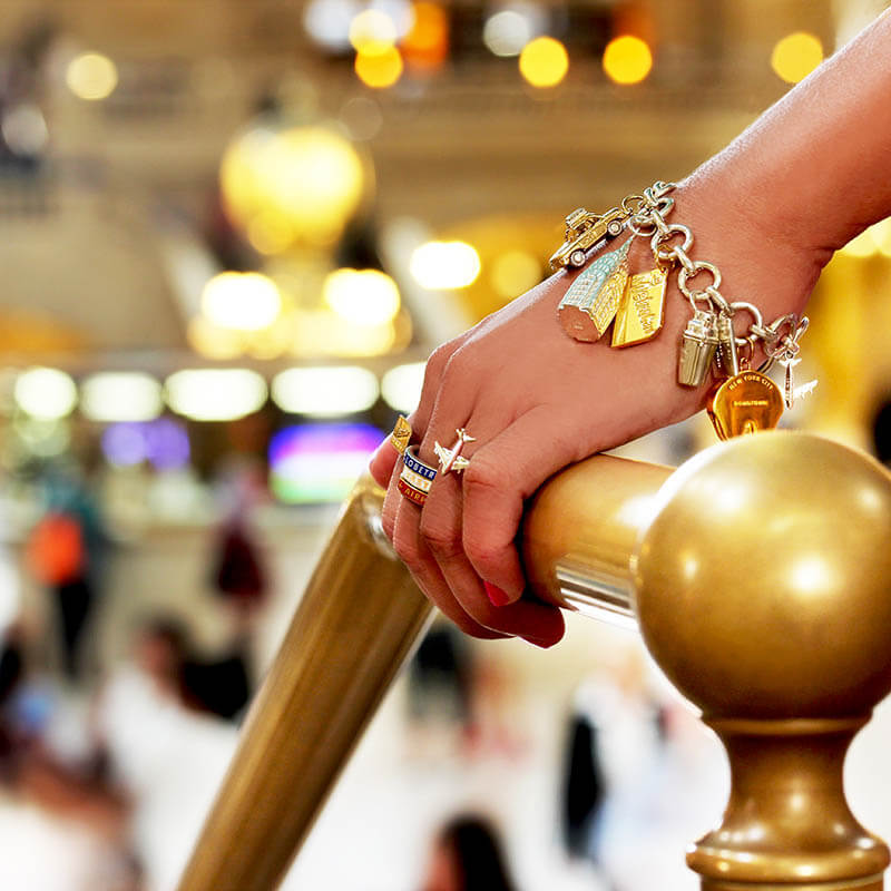 Hand wearing a New York charm bracelet with all New York Charms including an NYC taxi charm, a MetroCard charm, a Classic Manhattan cocktail shaker charm, and a Chrysler Building Charm. The hand is holding a gold rail and the main room in grand central is in the background