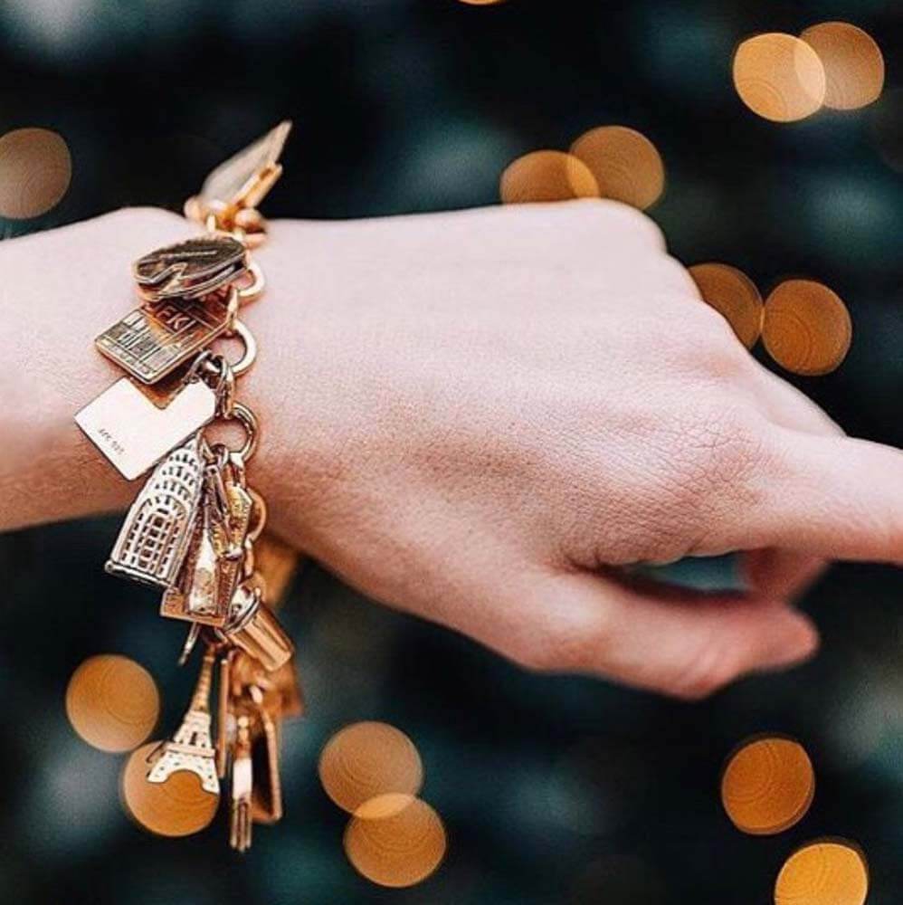 Gold Infinity Link Charm Bracelet from JET SET CANDY shown on a hand and featuring a variety of travel-inspired charms including a gold Eiffel Tower charm and a  gold  Chrysler Building charm. Background is dark green with yellow bokeh flairs